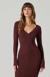 A woman is wearing a fitted, long-sleeved ribbed dress in a deep burgundy color. The dress features a V-neckline and vertical ribbing throughout. She accessorizes with a gold chain necklace and a ring on her left hand.